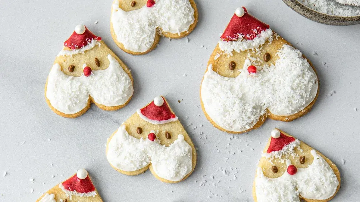 Mehrere herzförmige Kekse, als Weihnachtsmänner dekoriert, mit roten Hüten, weißen Kokos-Bärten und roten Nasen.