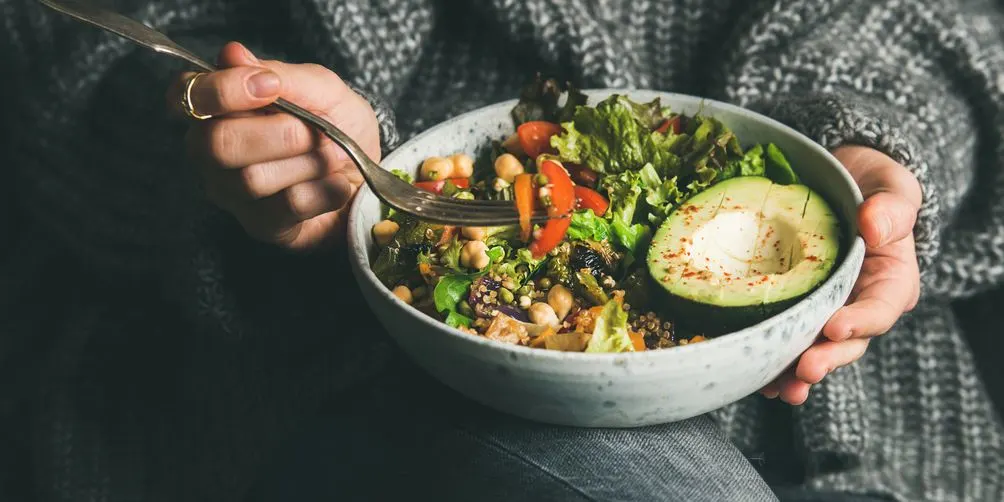 Person in grauer Strickjacke hält eine Schüssel mit Salat, Avocado, Kichererbsen, Tomaten und buntem Gemüse in den Händen und isst mit einer Gabel.