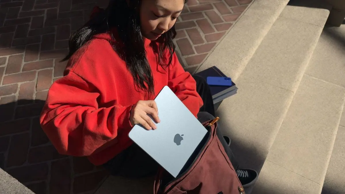 Frau in rotem Pullover sitzt auf einer Steintreppe, hält ein silbernes MacBook und steckt es in eine braune Tasche.