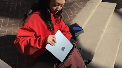 Frau in rotem Pullover sitzt auf einer Steintreppe, hält ein silbernes MacBook und steckt es in eine braune Tasche.