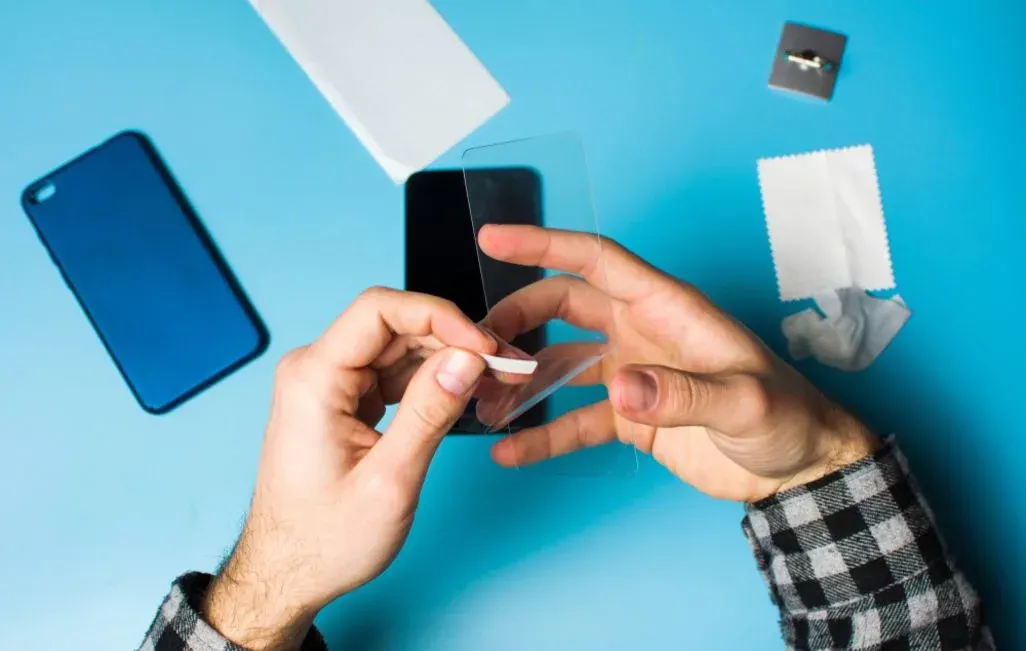 Hände ziehen Schutzfolie von einem Display-Schutzglas ab. Auf blauem Untergrund liegen ein Smartphone, eine Hülle und Reinigungstücher.