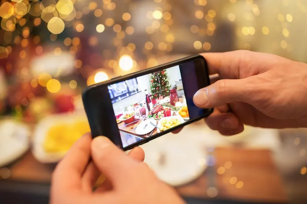 Hände halten ein Smartphone, das einen festlich gedeckten Tisch mit Braten, Salaten und einem Weihnachtsbaum zeigt. Goldene Bokeh-Lichter im Hintergrund.