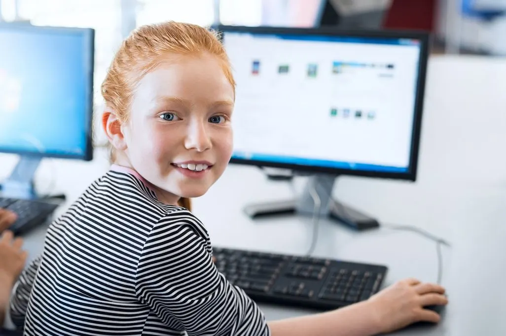 Ein lächelndes Kind in gestreiftem Shirt nutzt eine Maus am Computer. Zwei Monitore sind auf einem hellen Tisch zu sehen.