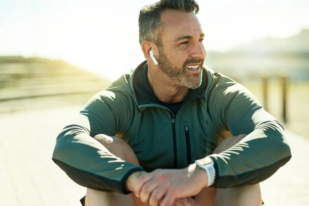 Mann in grüner Sportjacke sitzt draußen mit Kopfhörern und Smartwatch am Handgelenk.