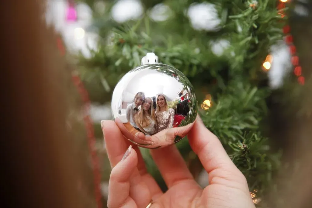 Hand hält glänzende silberne Kugel mit Reflexion von drei Personen und Raum. Unscharfer Weihnachtsbaum mit Lichtern und roten Girlanden.