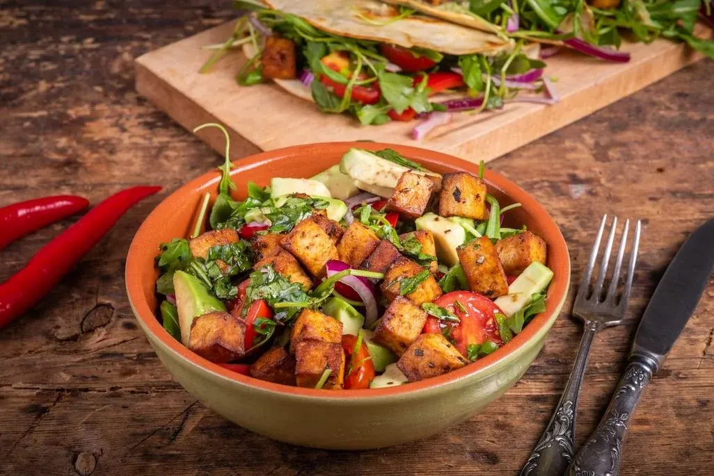 Bunte Salatschüssel mit braunem Tofu, grüner Avocado, roten Tomaten und Zwiebeln auf Holztisch. Rote Chilis, Besteck, Tacos auf Brett im Hintergrund.