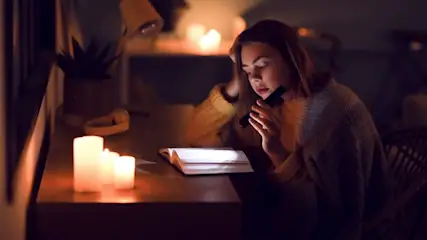 Person in gelbem Pullover liest Buch mit Taschenlampe. Drei Kerzen und eine Pflanze auf dunklem Holztisch im gedimmten Raum.