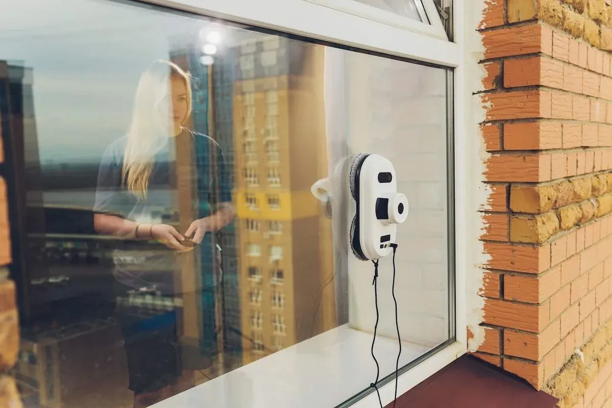 Fensterroboter sitzt an der Außenseite eines Fenster auf der Scheibe. Durch diese sieht man durch die Spiegelungen eine Frau, die den Roboter per Fernbedienung steuert.