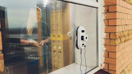 Fensterroboter sitzt an der Außenseite eines Fenster auf der Scheibe. Durch diese sieht man durch die Spiegelungen eine Frau, die den Roboter per Fernbedienung steuert.