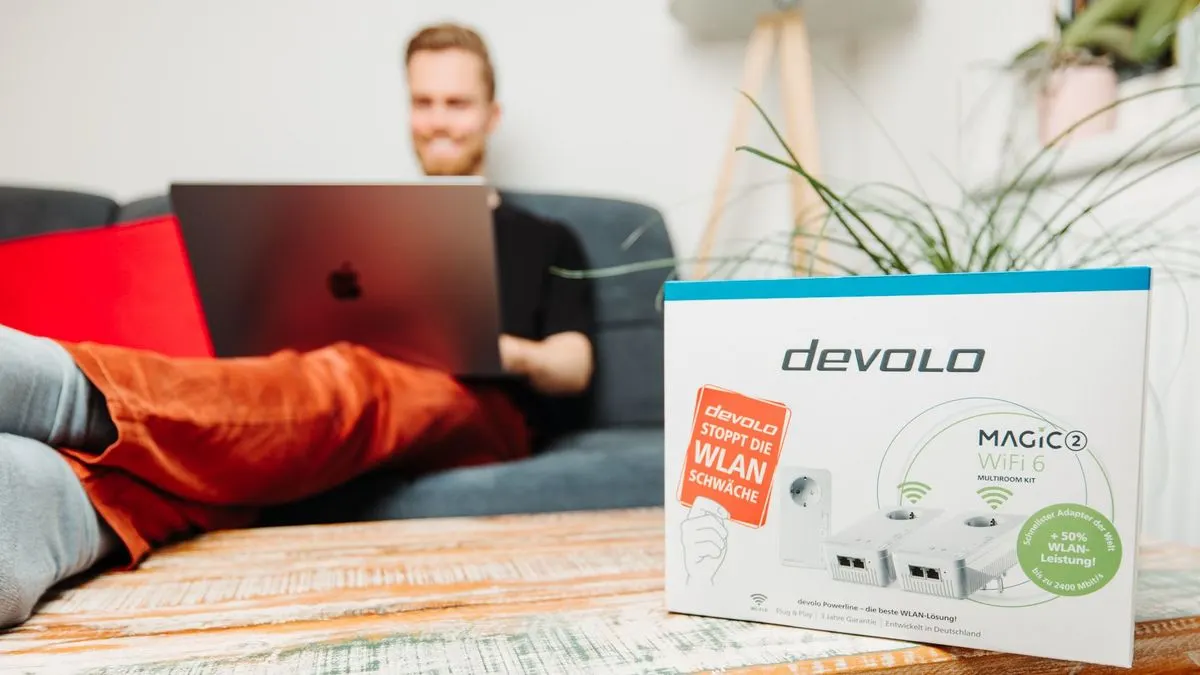 Person sitzt mit einem Laptop auf dem Sofa, im Vordergrund steht eine Produktverpackung von devolo Magic 2 WiFi 6.
