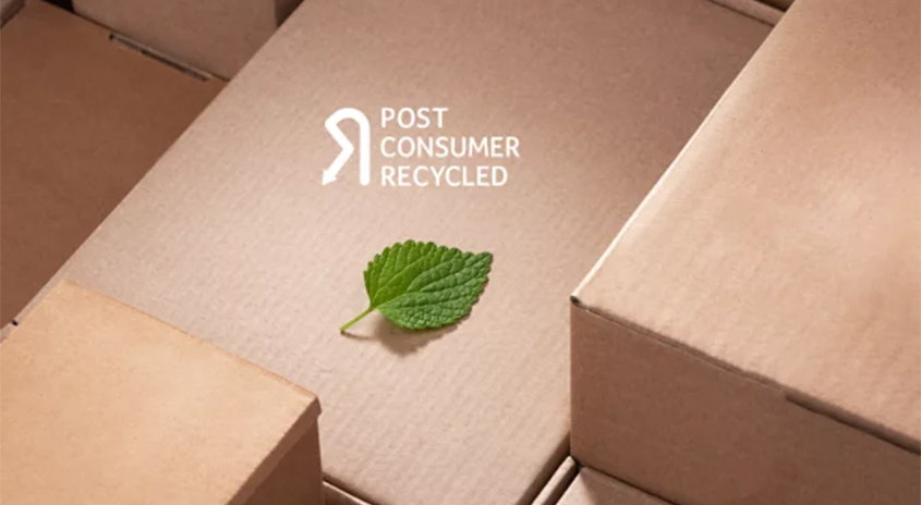 Auf braunen Kartons liegt ein grünes Blatt. Text "Post Consumer Recycled" in Weiß. Fokus auf Nachhaltigkeit und Recycling.