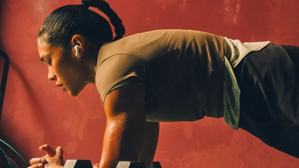 Eine Person in braunem Shirt und dunklen Shorts macht einen Plank auf einer Hantel. Schweiß ist sichtbar, weiße Apple Air Pods Pro 3 Ohrstöpsel im Ohr, roter Hintergrund.