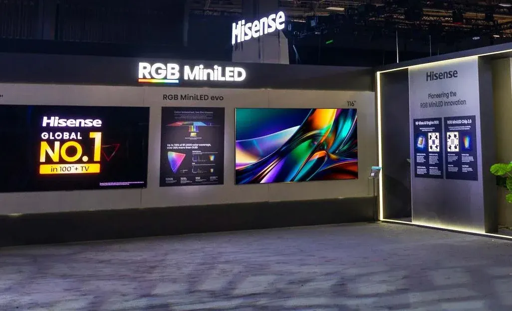 Dunkel beleuchteter Messestand mit prominentem „RGB MiniLED“-Schriftzug und Hisense-Logo. Mehrere große Bildschirme an einer Wand zeigen links eine „GLOBAL NO.1“-Anzeige, in der Mitte ein Informationsposter und rechts ein farbenfrohes TV-Bild; rechts daneben sind beleuchtete Infotafeln zur RGB MiniLED-Technologie zu sehen.