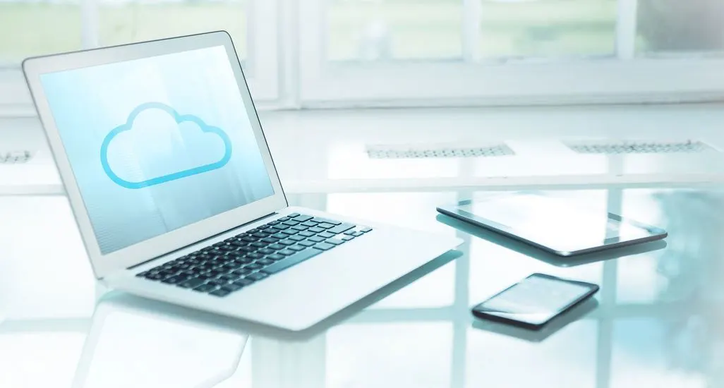 Laptop mit Cloud-Symbol auf Bildschirm neben Tablet und Smartphone auf Glastisch.