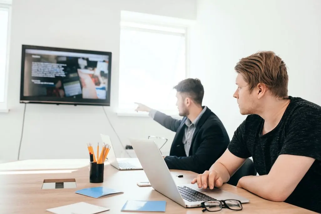 Zwei Maenner sitzen am Tisch mit Laptops und schauen auf großen Bildschirm an der Wand.