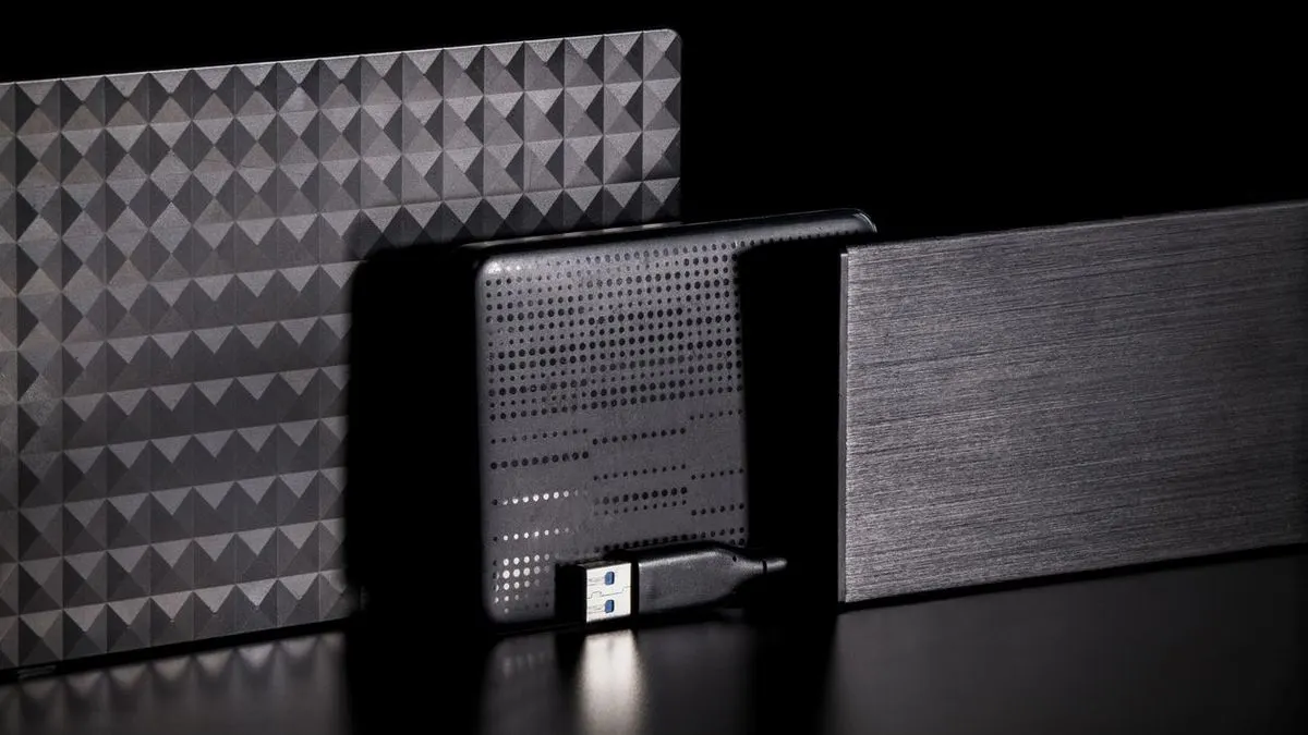Drei externe Festplatten mit unterschiedlichen Oberflächenstrukturen stehen nebeneinander auf einem schwarzen Tisch. Davor liegt ein USB-Kabel.