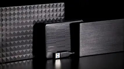 Drei externe Festplatten mit unterschiedlichen Oberflächenstrukturen stehen nebeneinander auf einem schwarzen Tisch. Davor liegt ein USB-Kabel.