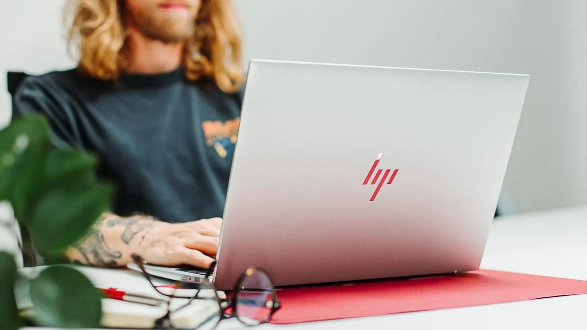 Ein Mann mit langen blonden Haaren arbeitet an einem silbernen HP Laptop an einem Schreibtisch.