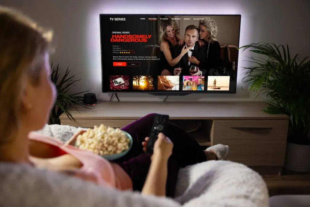 Eine Person sitzt auf einem Sofa, hält Popcorn und Fernbedienung. Ein beleuchteter Fernseher zeigt eine Streaming-Serie über einen Mann und zwei Frauen, mit Pflanzen auf dem TV-Möbel.