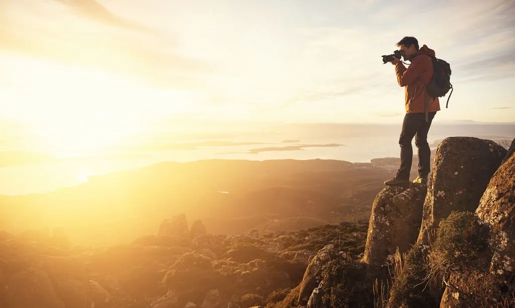 Person mit Rucksack fotografiert Sonnenuntergang auf einem Felsen mit weitem Ausblick