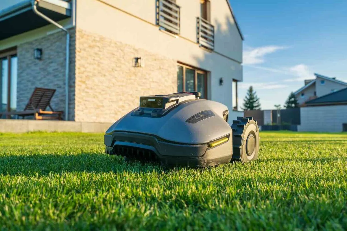  Rasenmähroboter fährt über einen dicht bewachsenen, grünen Rasen vor einem modernen Einfamilienhaus bei Sonnenschein.