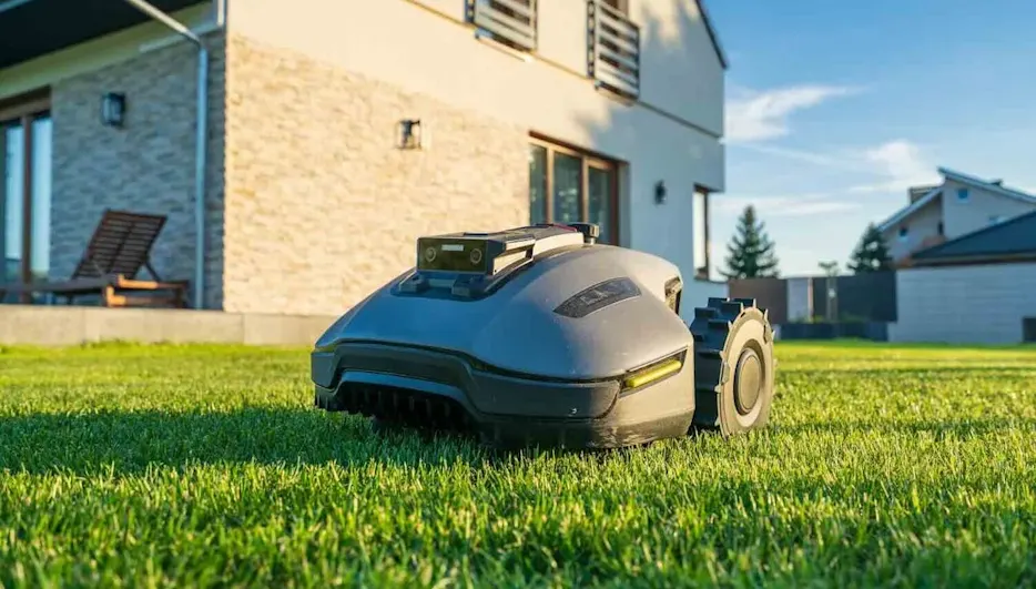 Rasenmähroboter fährt über einen dicht bewachsenen, grünen Rasen vor einem modernen Einfamilienhaus bei Sonnenschein.