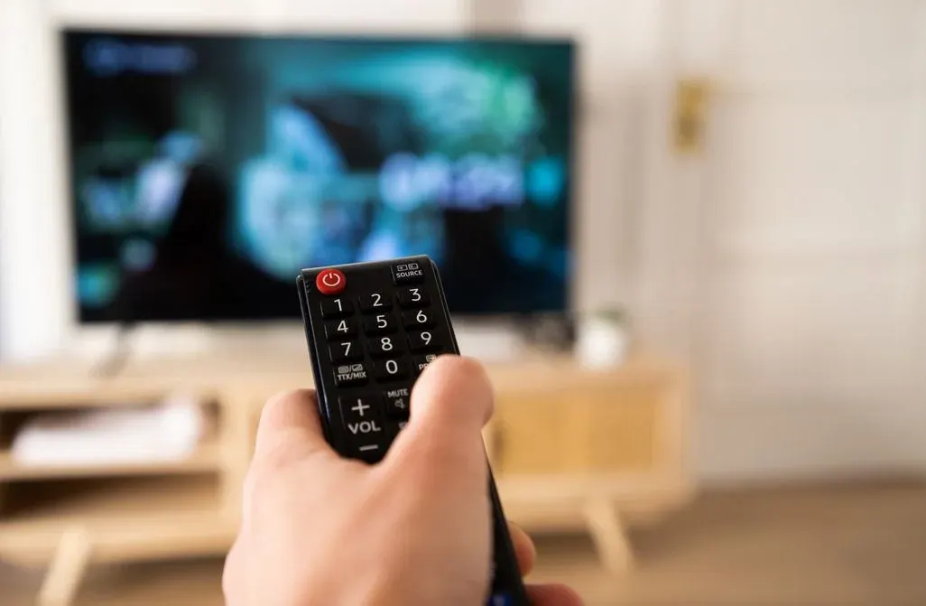 eine Hand hält eine Fernbedienung, im Hintergrund sieht man einen Fernseher