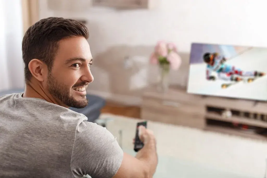  Mann sitzt lächelnd im Wohnzimmer mit Fernbedienung in der Hand und schaut auf einen Fernseher, auf dem ein Skirennen läuft.