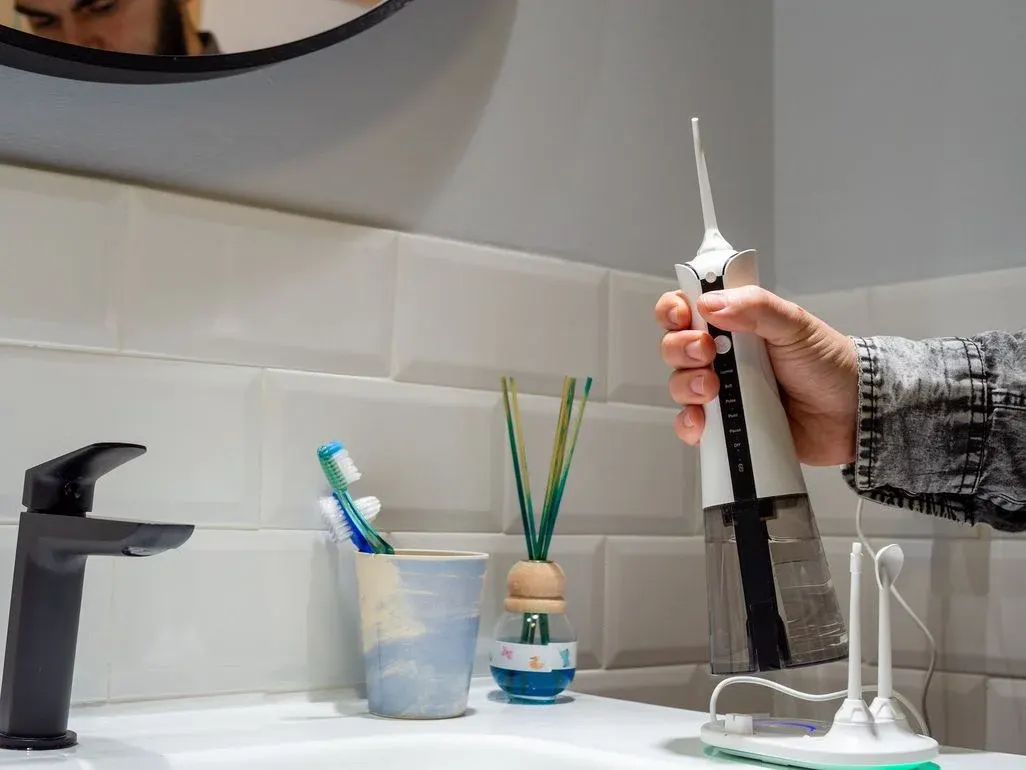 Hand hält weiße Munddusche über Waschbecken mit schwarzem Wasserhahn. Zahnbürsten, Diffusor, Ladestation auf Theke. Person im Spiegel.