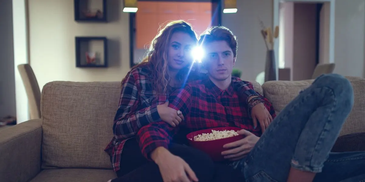 Zwei Personen auf hellem Sofa in karierten Hemden. Mann hält rote Popcornschale. Heller Lichtschein. Im Hintergrund Wandregale und Tür.