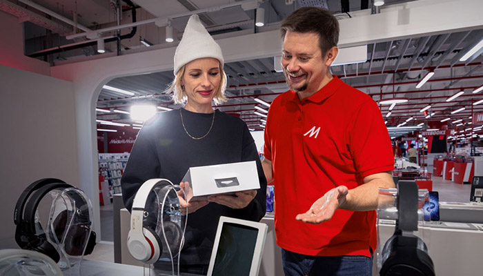 In einem Elektronikgeschäft sehen wir eine Frau mit Mütze, die Kopfhörer und eine Box hält, neben einem Mann in rotem Hemd.