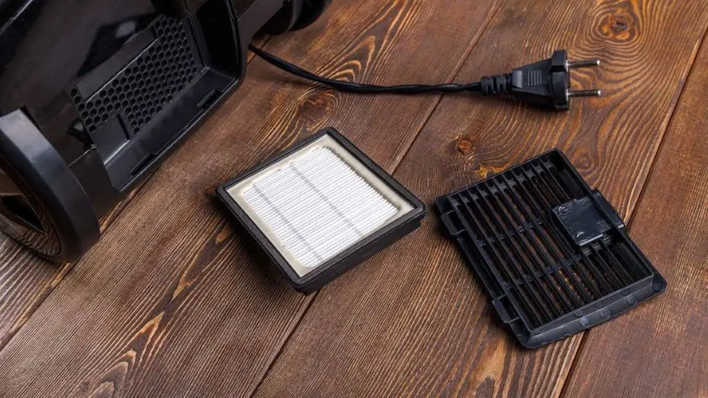 Ein schwarzer Staubsauger mit geöffnetem Fach, daneben ein weißer Filter und schwarzes Gitter. Ein schwarzer Stecker liegt auf braunem Holzboden.