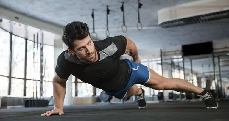 Mann in schwarzem Shirt und blauen Shorts macht einen einarmigen Liegestütz auf dunklem Gymnastikboden. Im Hintergrund Fenster und Turnringe.