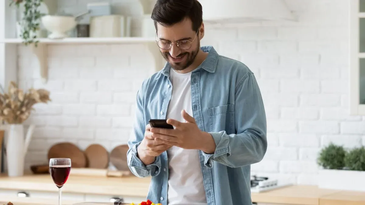 Lächelnder Mann in Jeanshemd und Brille hält Smartphone. Rotweinglas, gehacktes Gemüse und Messer auf einer Fläche.