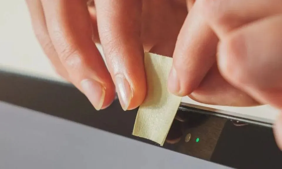 Eine Hand klebt ein Stueck Klebeband auf die Kamera eines Laptops