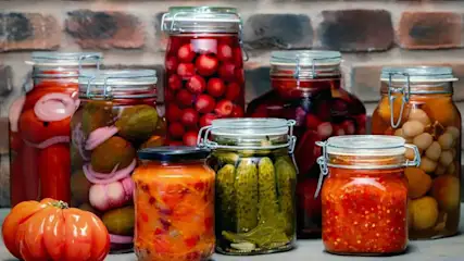 Mehrere Einmachgläser mit eingelegtem Gemüse – Gurken, Tomaten, Paprika und Zwiebeln – vor einer Backsteinwand, daneben eine frische Tomate.