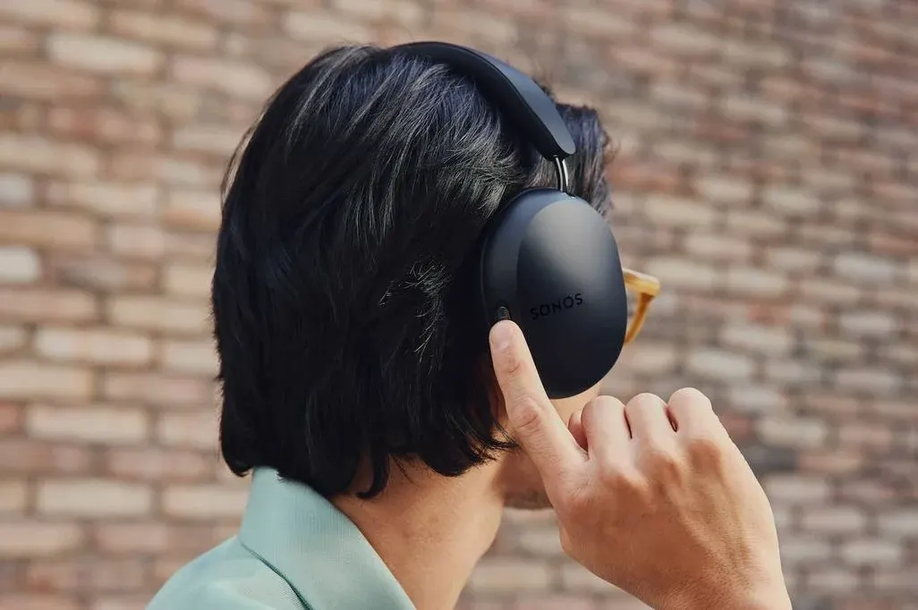 Person mit schwarzen Sonos-Kopfhörern, die von hinten zu sehen ist. Sie trägt einen hellblauen Pullover und bedient die Kopfhörer mit einem Fingertipp. Im Hintergrund ist eine Backsteinmauer sichtbar.