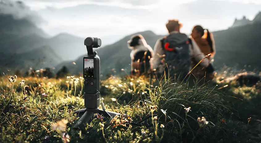 Eine kleine Kamera auf einem Stativ steht in einem Grasfeld mit Bergen im Hintergrund.