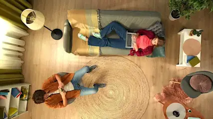 Wohnzimmer von oben: Person liegt mit Laptop entspannt auf dem Sofa, zweite Person sitzt davor. Heller Holzboden, runder Teppich, Pflanzen.