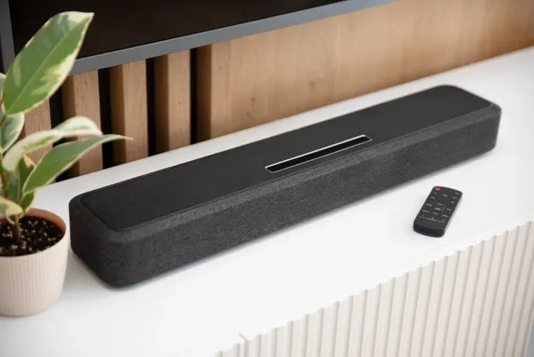 Schwarze Soundbar mit grauem Stoff und silbernem Schlitz, daneben schwarze Fernbedienung und Topfpflanze auf weißer Oberfläche.