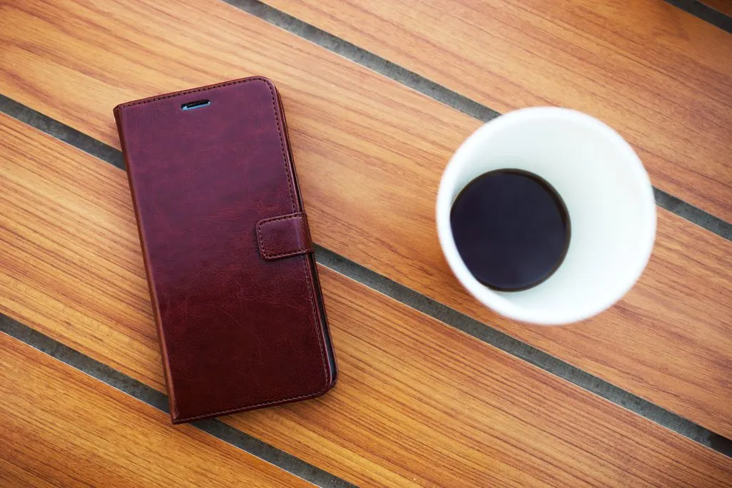 Braune Smartphone-Huelle liegt neben einem Becher Kaffee auf einem Holztisch