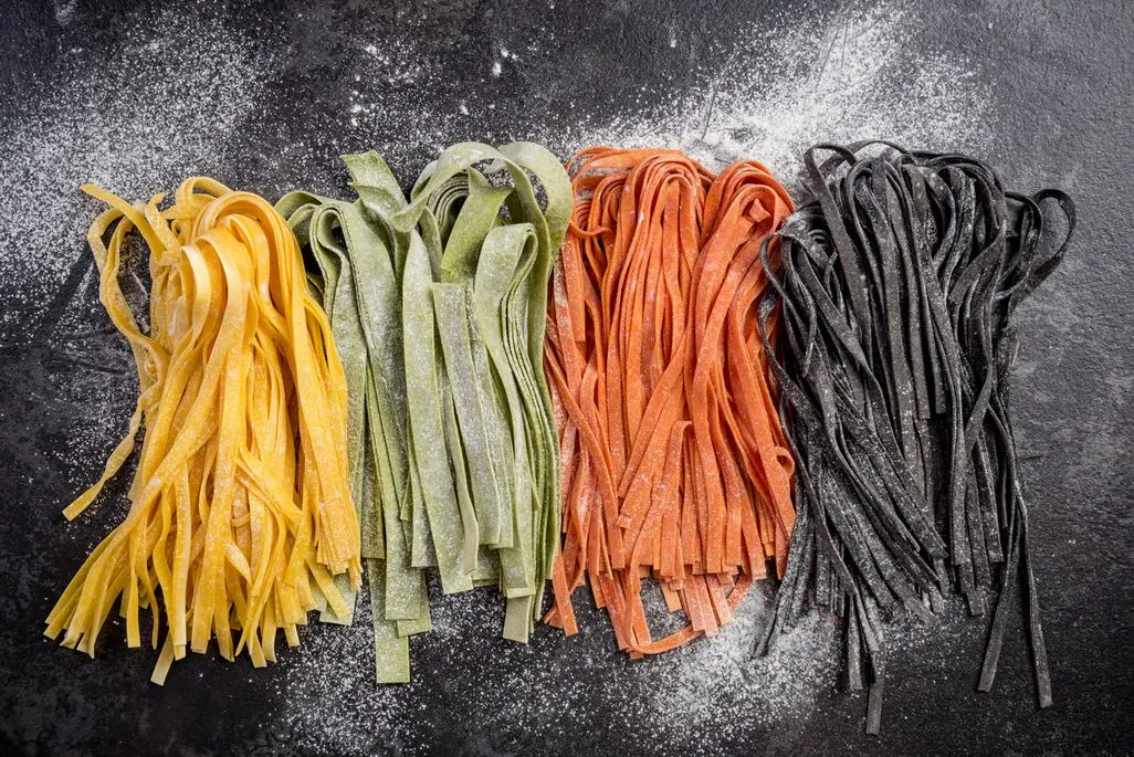 Vier Bündel roher Tagliatelle in Gelb, Grün, Orange und Schwarz, bestäubt mit Mehl, auf dunklem Untergrund.