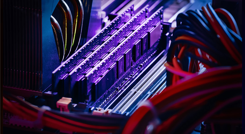 Nahaufnahme von Computerkomponenten: RAM-Module mit Kühlkörpern, bunte Kabel. Violette Beleuchtung betont die Details.