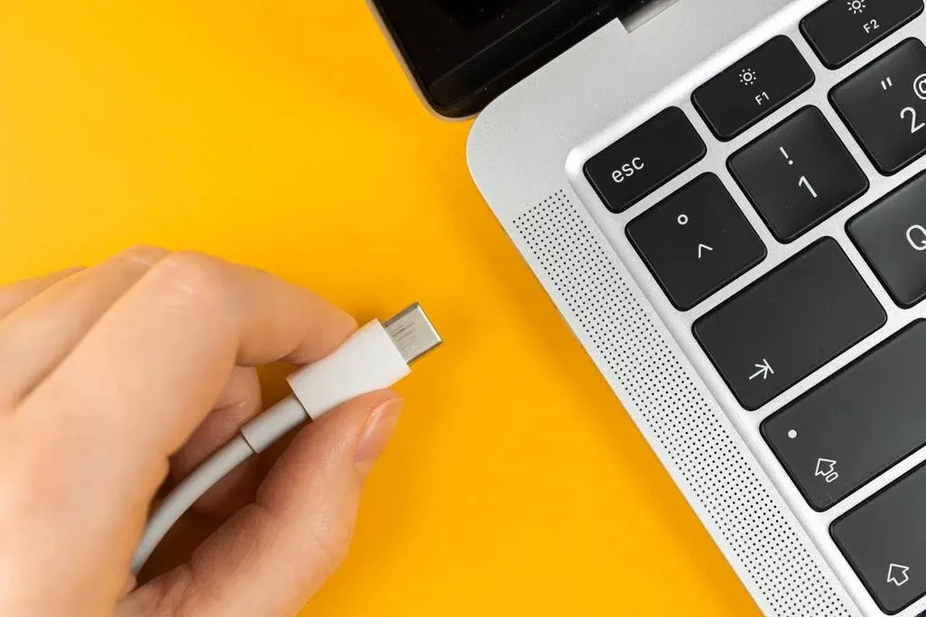 Eine Hand hält ein weißes USB-C-Kabel nahe einer silbernen Laptoptastatur mit schwarzen Tasten.
