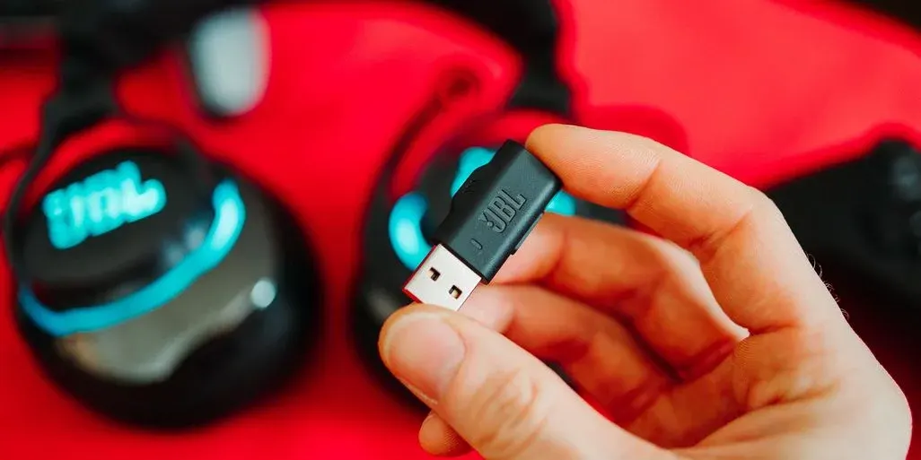 Eine Hand hält einen schwarzen JBL USB-Dongle mit rotem Stecker. Im Hintergrund ist ein schwarzer Kopfhörer mit blauem JBL-Logo.