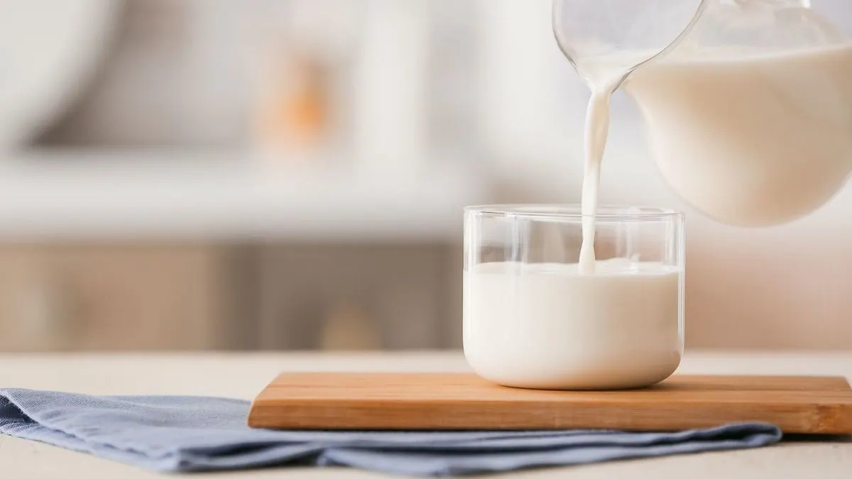 Milch wird aus einem Krug in ein Glas gegossen, das auf einem Holzbrett mit blauem Tuch steht. Heller, unscharfer Hintergrund.