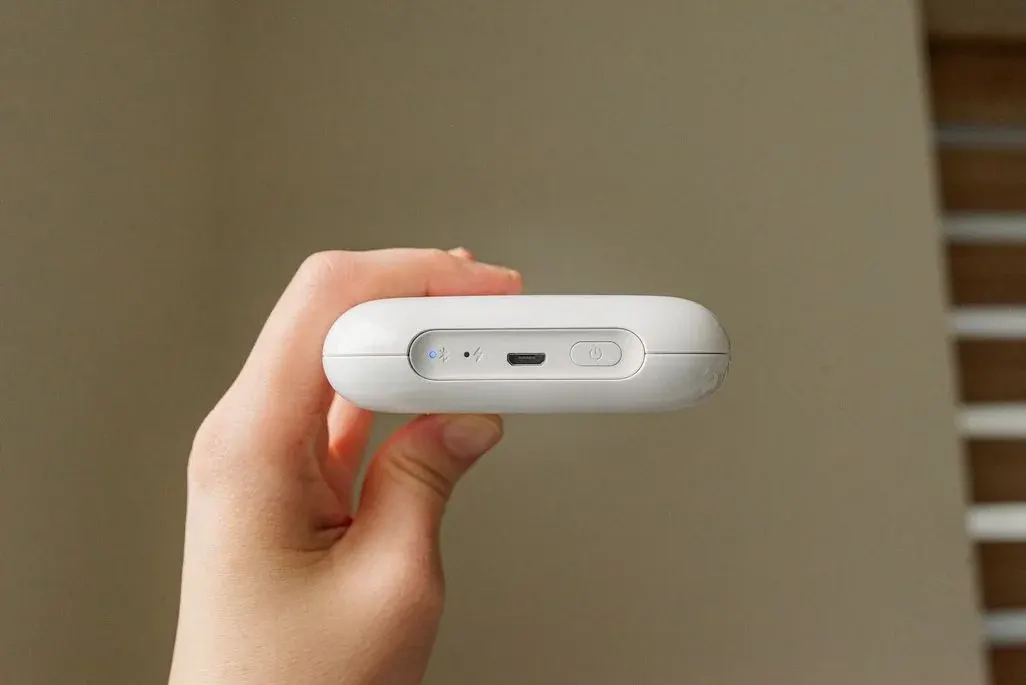 Hand hält weißes tragbares Gerät mit Bluetooth-Symbol, USB-C-Anschluss und Power-Button – kompaktes technisches Zubehör vor neutralem beigem Hintergrund.