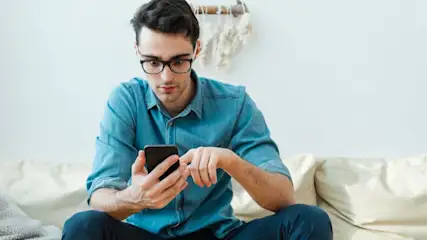 Mann mit Brille und blauem Hemd sitzt auf heller Couch, tippt auf schwarzem Smartphone. Weiße Wand mit heller Deko im Hintergrund.