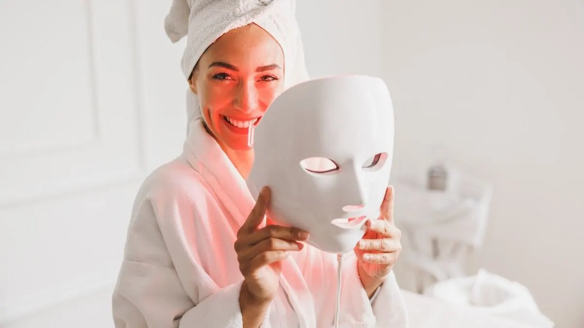 Eine Person mit weißem Handtuch und Bademantel hält eine weiße LED-Maske mit rotem Licht vor ihr Gesicht in einem hellen Raum.