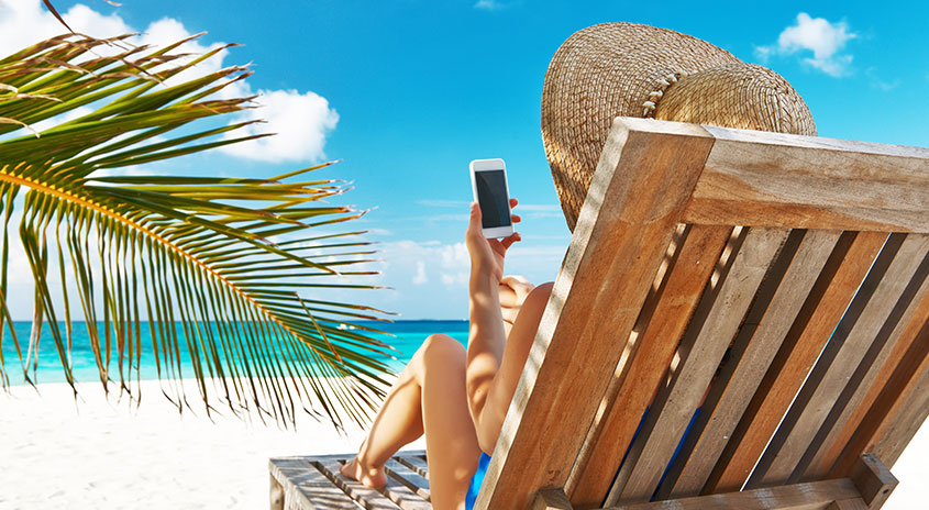Eine Person sitzt auf einem Holzstuhl am Strand und hält ein Smartphone vor blauem Himmel und Meer.